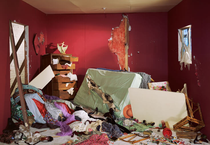 A bedroom in a state of disarray, the ground covered with clothing and a shredded mattress.