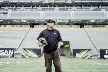 A man stands at the centre of a football field cradling a football helmet under his arm and wearing a polo shirt and baseball cap.