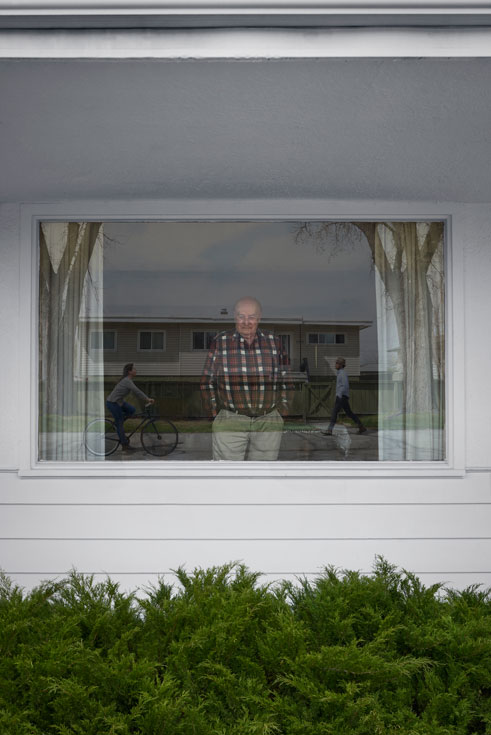 An older man looks out the window. Reflected in the glass are a cyclist and a pedestrian walking outside.