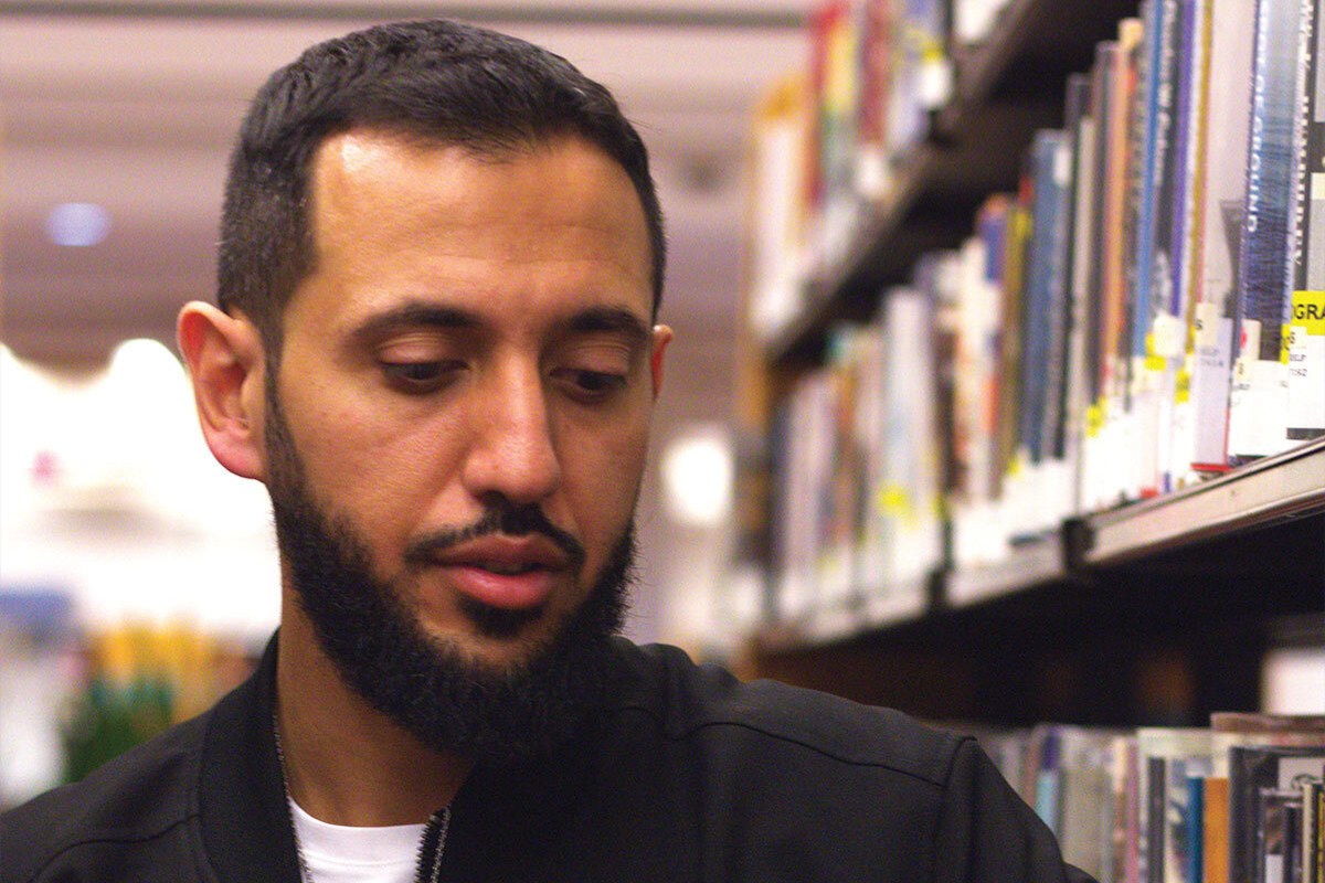 In front of a library bookcase, a man wearing a black jacket over a white T-shirt folds his hands.