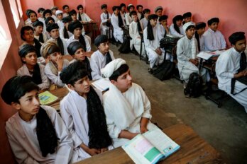 Afghan boys, wearing long white robes, sitting on benches with a few school books in front of them