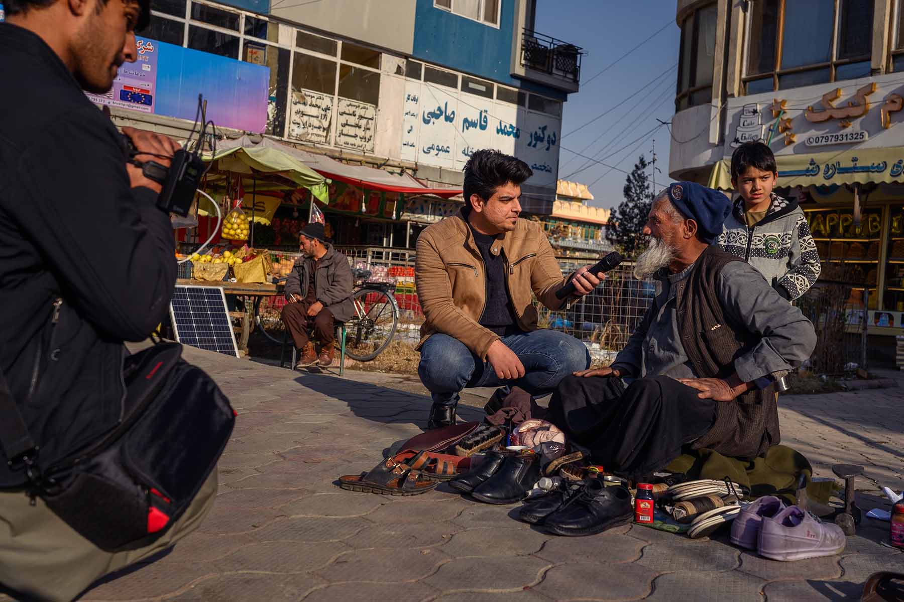 An online producer talks to an older man selling shoes on the street in Kabul.
