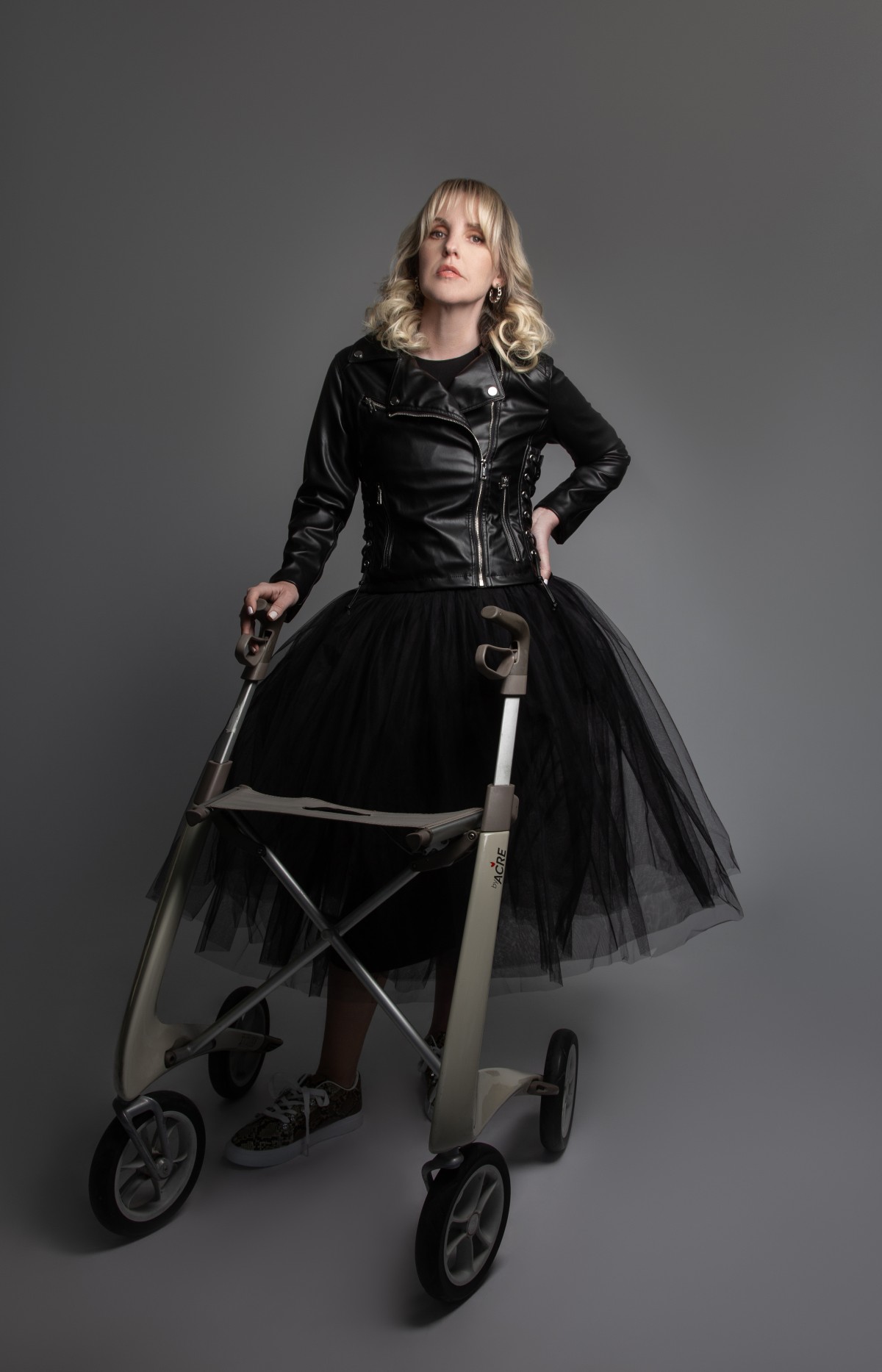 A photo of a blonde woman in a leather jacket and black tulle skirt holding the handles of a mobility device