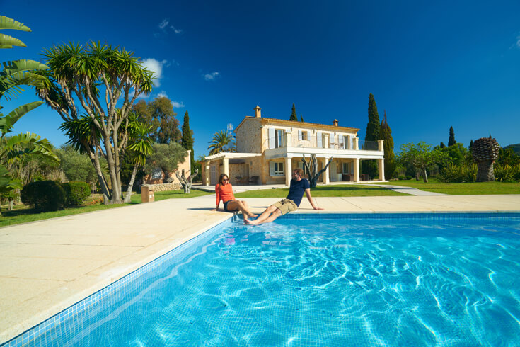A man and a woman sit on the edge of a large pool with their legs in the water; behind them is an elaborate villa against a backdrop of trees and a blue sky.