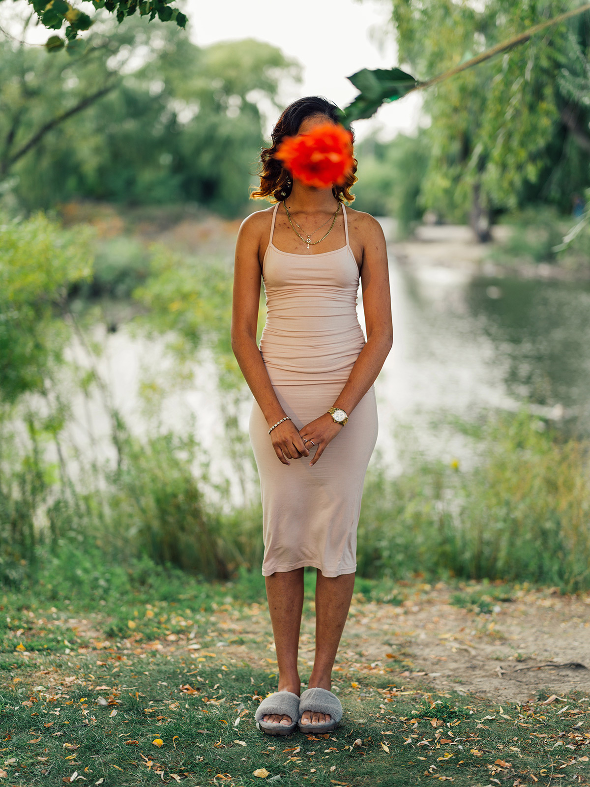 Photo of a teenage girl wearing a spaghetti-strap dress and fuzzy sandals. She is standing outside near a lake with green trees and bushes visible. A single out of focus red dhalia hangs down from the edge of her frame, blocking her face from view.