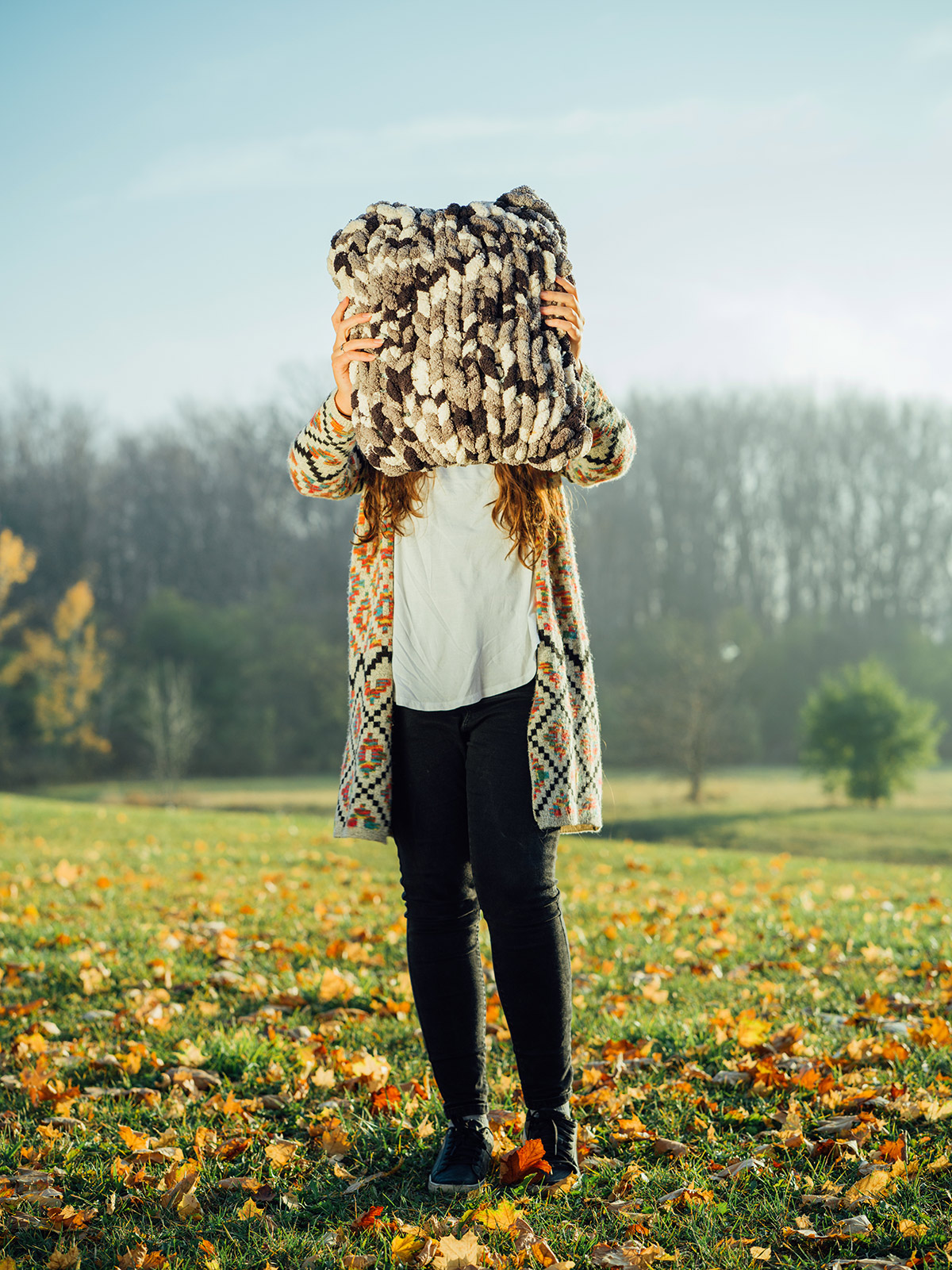 Photo of a teenage girl wearing a long sweater and black pants. She is standing in a field with colourful fallen leaves on the ground. She is holding a large crocheted pillow in front of her face.