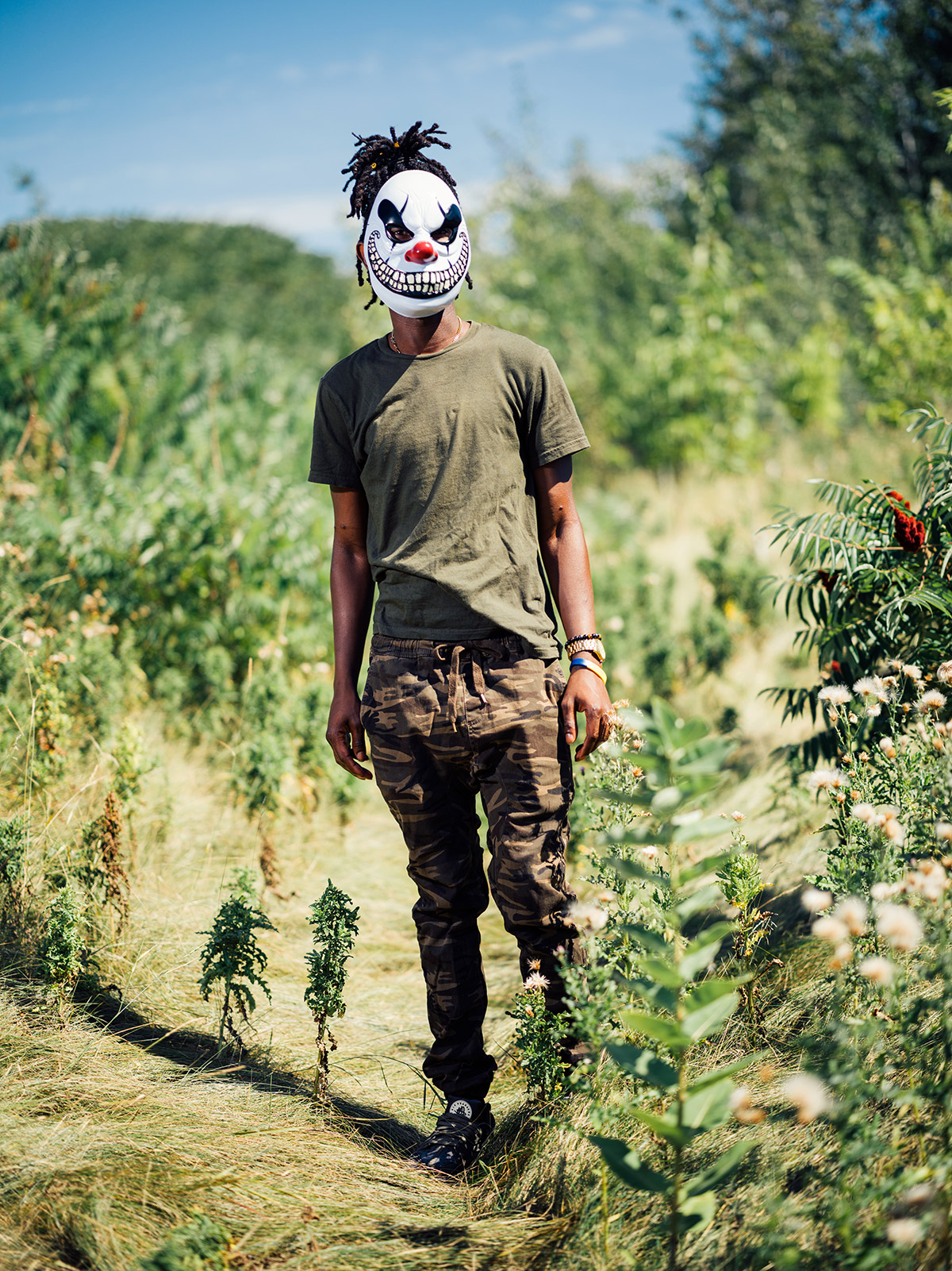 Photo of a teenage boy standing among trees and bushes wearing a green t-shirt and camo pants. His hair is in dreadlocks and tied up on top of his head. He is wearing a clown mask with a wide, curling, toothy smile.