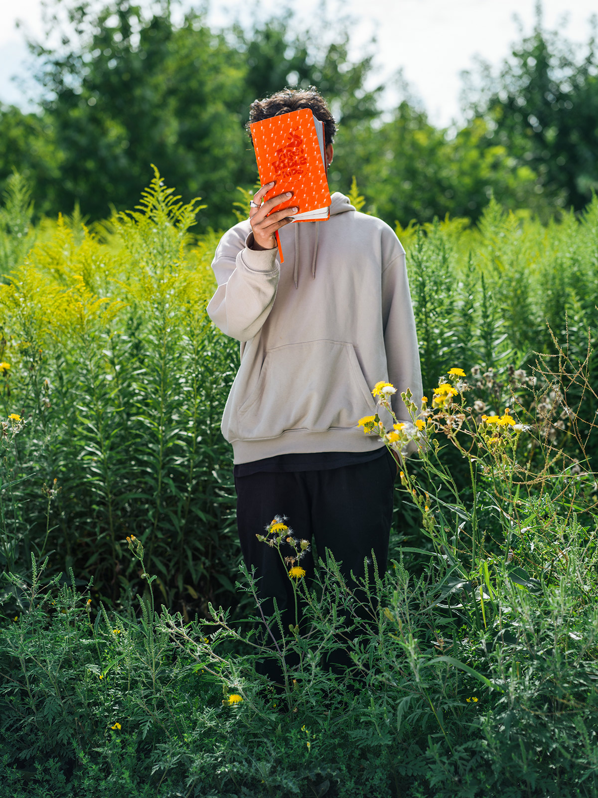 Photo of a teenage boy, wearing a grey hoodie, standing in a field of knee-height weeds. He is holding a bright orange book in front of his face, the cover reading 'Fire Bible for Kids.'