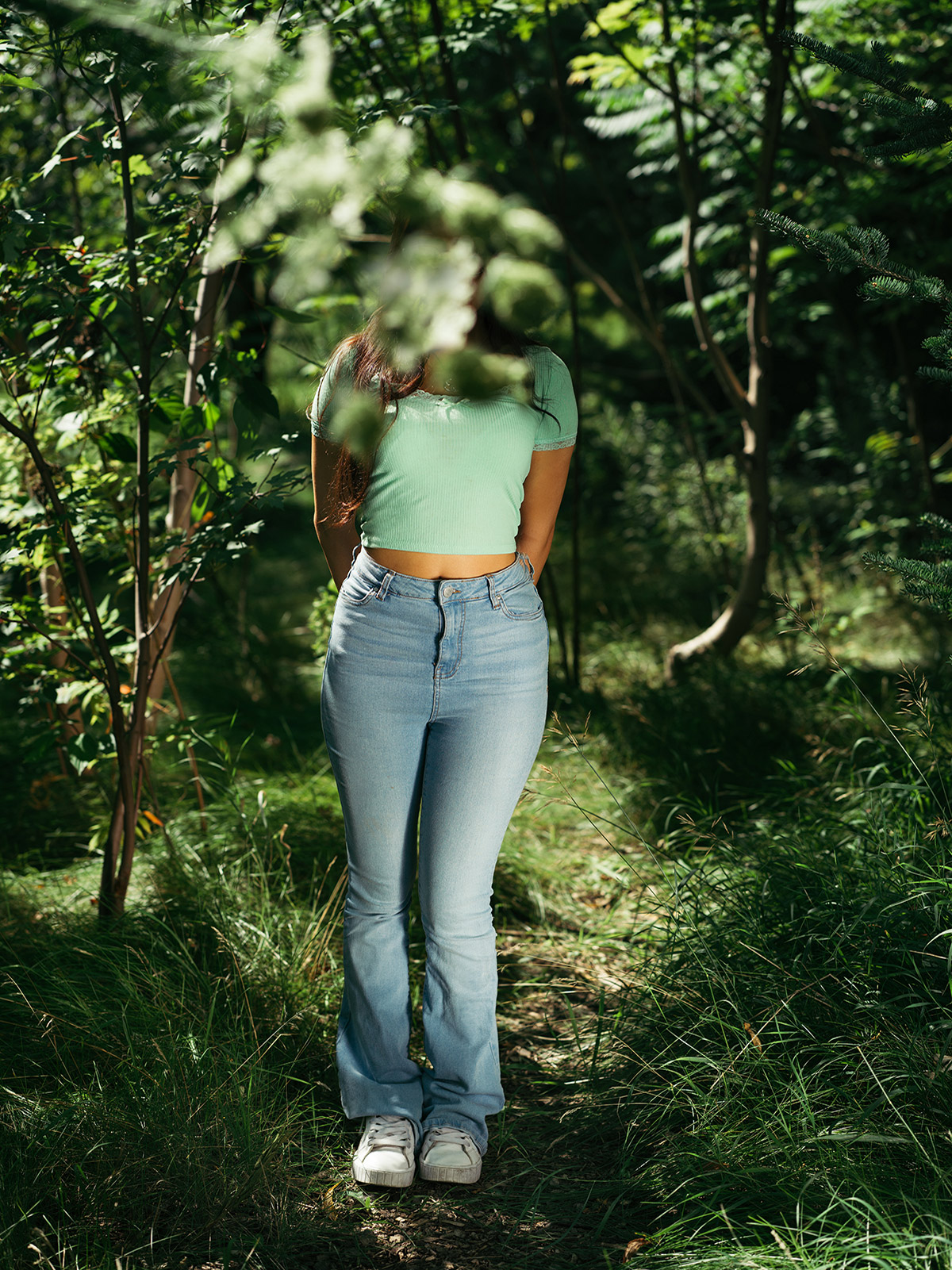 Photo of a teenage girl wearing a green t-shirt and light-wash jeans. She is standing in a forest and a tree branch hangs down from the edge of the frame, blocking her face from view.