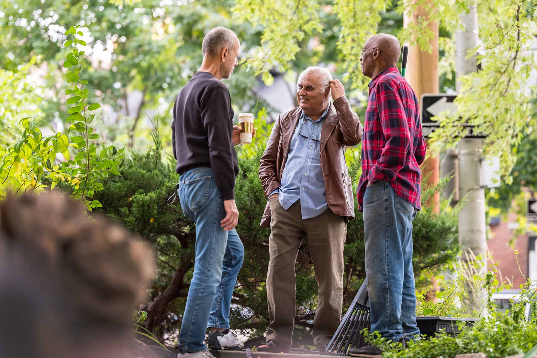 Three men, one holding a coffee, another leaning on a rake, chat together outside amid a street's trees and bushes.
