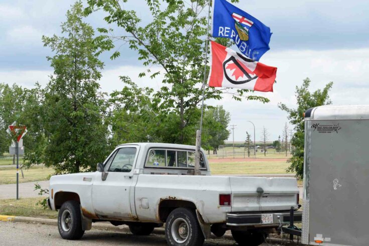 Photo of a white pickup truck driving and waving two flags on a pole: an upside-down Canadian flag with a crossed out maple leaf and an Alberta provincial flag with the word "Republic" added to it.