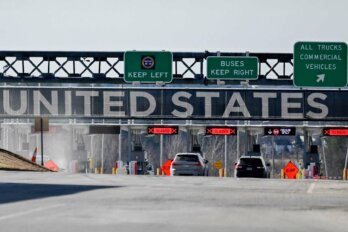 Photo shows two cars at a border crossing along a highway, with the words "United States" and three green signboards looming over them