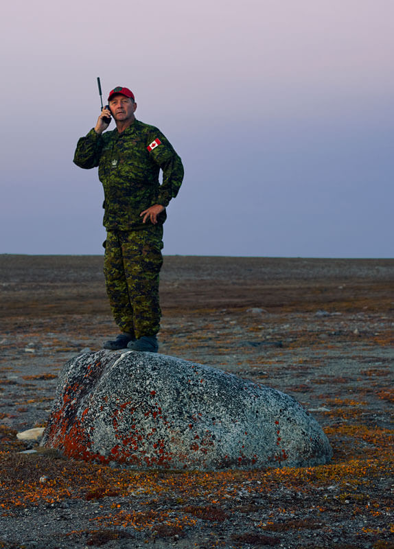 Man in a Canadian combat uniform holds a remote phone to his ear while standing on a rock.