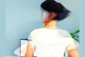 A woman sitting at a desk in front of a laptop and turning her head to the side, viewed from the back. Her face and shoulders are slightly blurred. She is wearing a white shirt and the wall in front of her is sky blue.