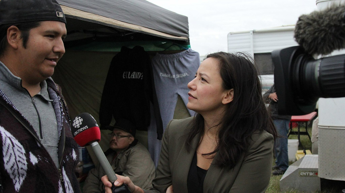 Photo of Connie Walker interviewing a man wearing a backwards baseball cap. She is holding a microphone with the logo of CBC News on it and a video camera lens is visible at the right edge of the photo.