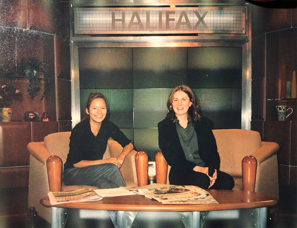 Photo of two young women, Connie Walker on the left and Danielle Stone on the right, sitting in chairs behind a coffee table in a TV studio. Above them is a sign reading 