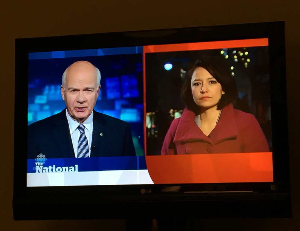 Photo of a TV airing CBC’s The National. Peter Mansbridge, a balding man in a suit and tie, is shown on the left side of the screen in front of a blue background. Connie Walker, wearing a red coat, is shown on the right side.