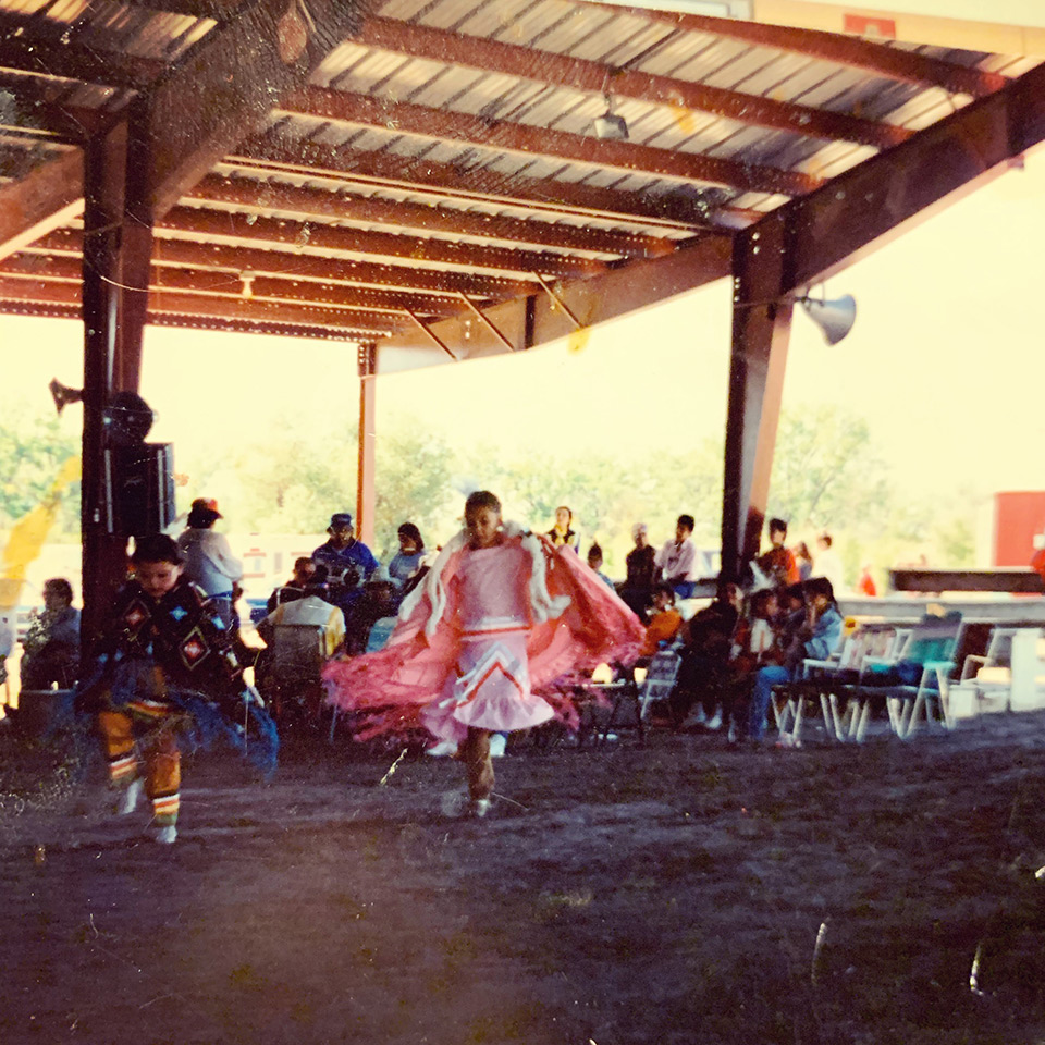 Photo of Connie Walker as a young girl wearing a pink shirt, skirt, and cape. She is dancing with others outdoors under a wooden roof. Groups of chairs and adults sit behind.