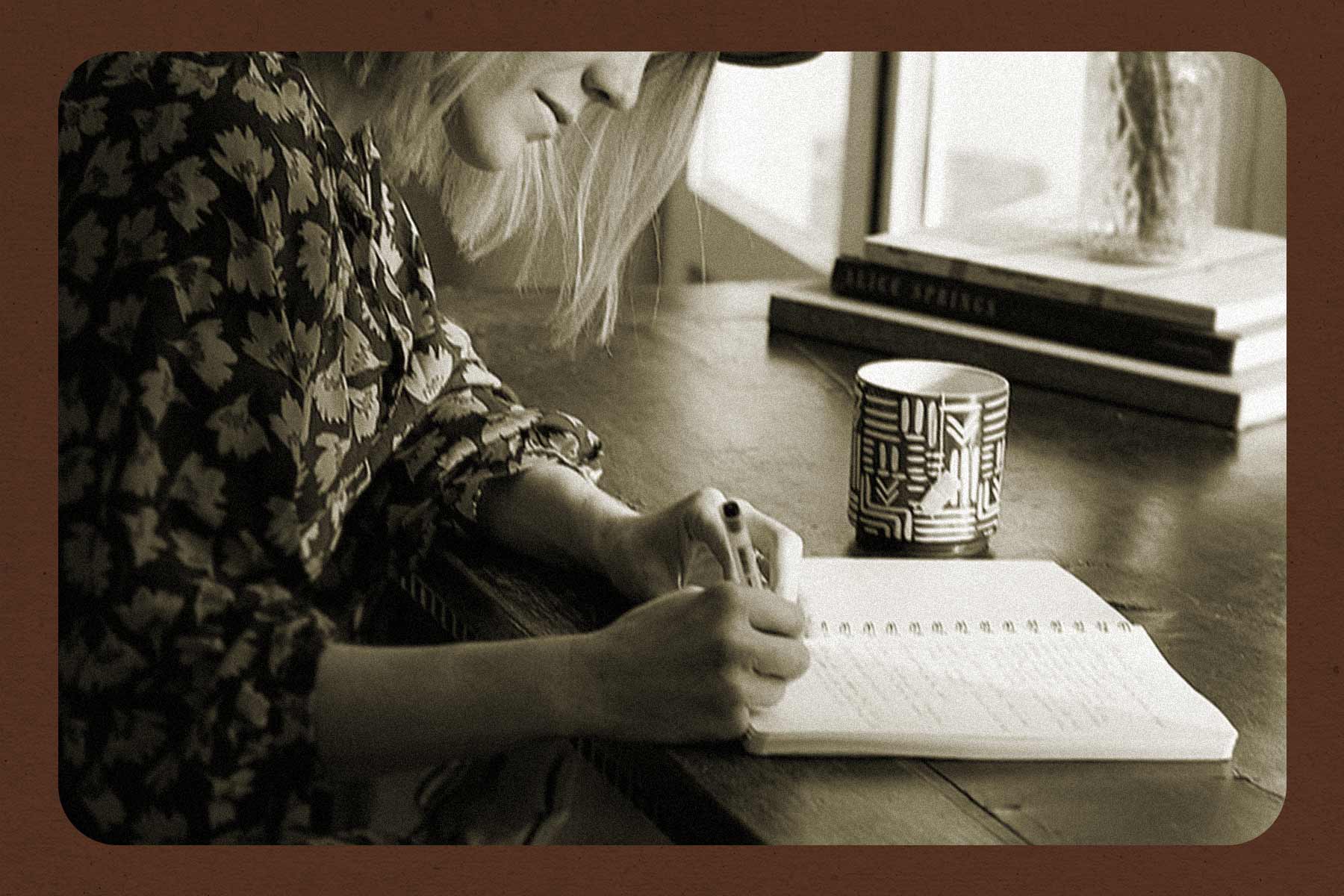 A black-and-white photo of a woman sitting at a desk and writing in a notebook. The image has a brown-coloured border around it.