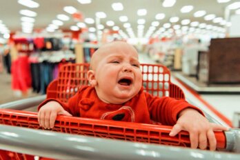 A photo of a baby crying while riding a shopping cart in a department store.