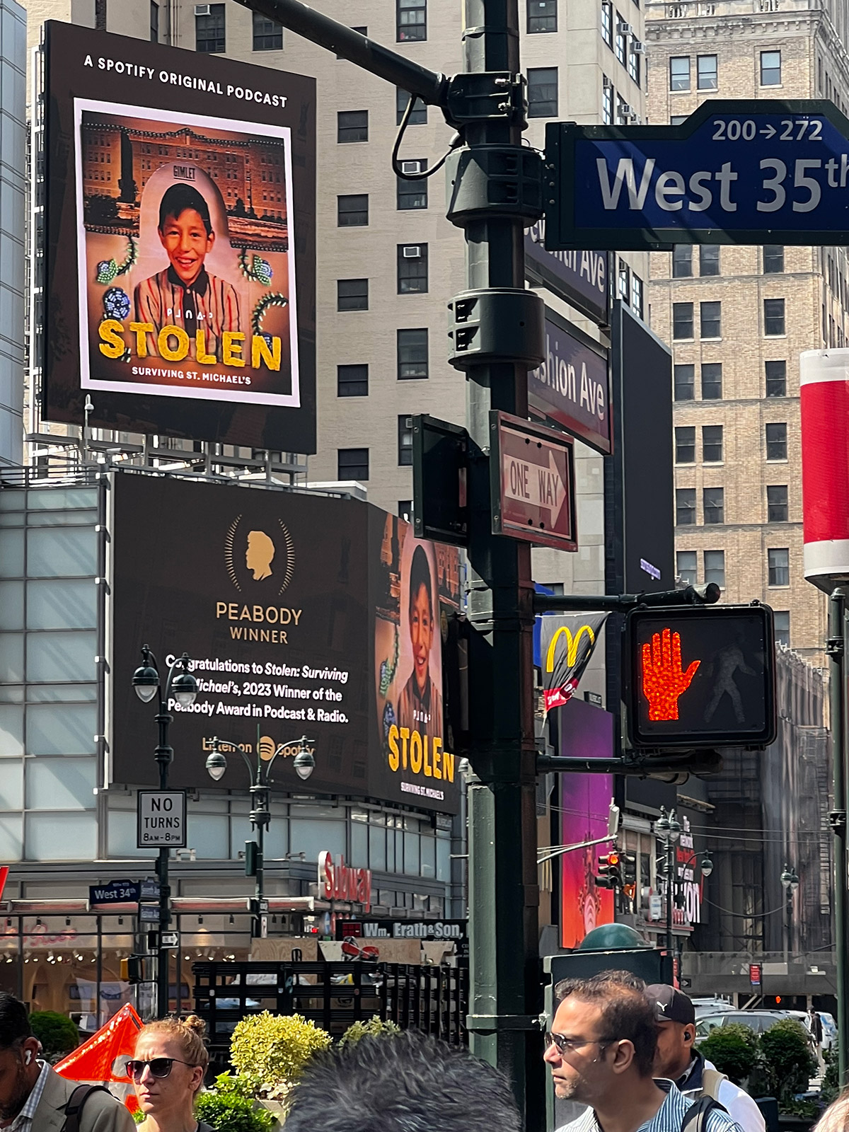 Photo of a busy street corner in New York City. Large electronic billboards show an image of a young smiling boy with the words 