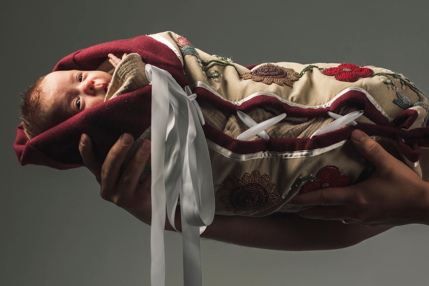 A baby held aloft against a grey background - they're swaddled in a quilted blanket