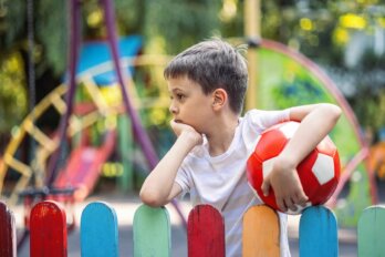 A bored boy, holding a red-and-white soccer ball, leans against a fence outside a playground