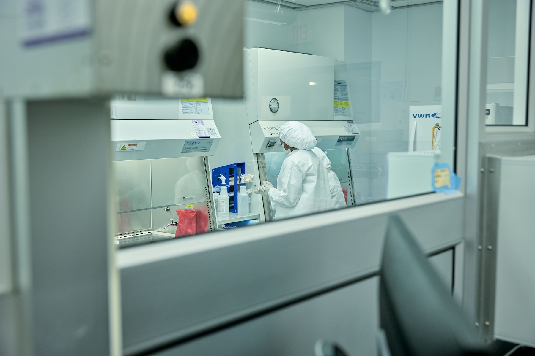 Photo looking through a window into a medical lab where a woman dressed in a white gown, hair covering, mask, and gloves is working.