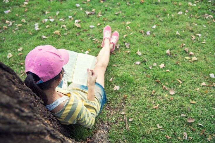 A girl with black hair reads a book while sitting under a tree.