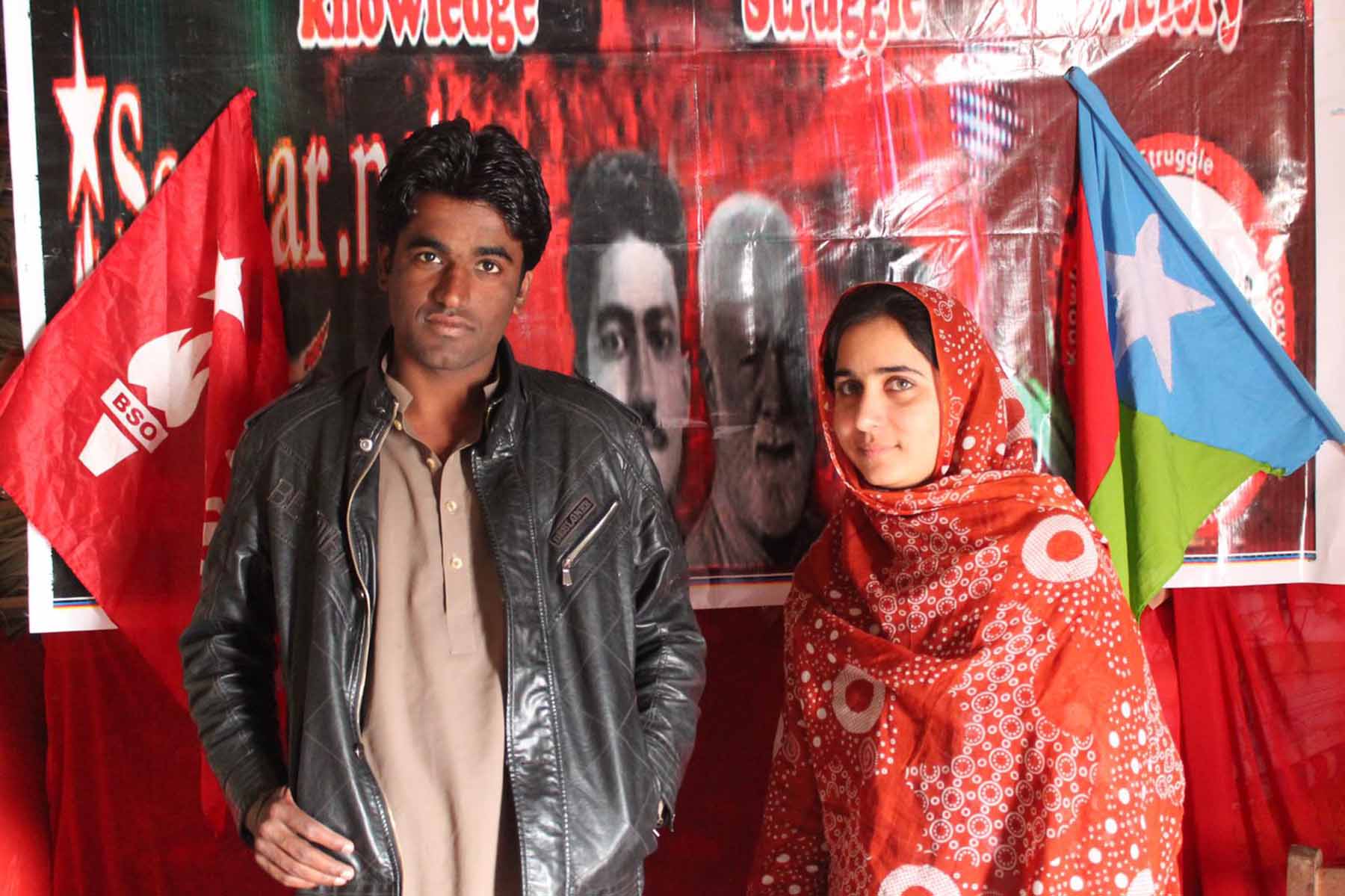 Lateef Johar, dressed in a leather jacket, and Karima Mehrab, dressed in red, stand in front of a red and black protest sign and blue, green and red Balochistan flag