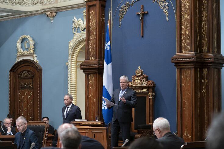 A man speaks on a stage at the Quebec Legislature with a crucifix over his head on the dark blue wall behind him.