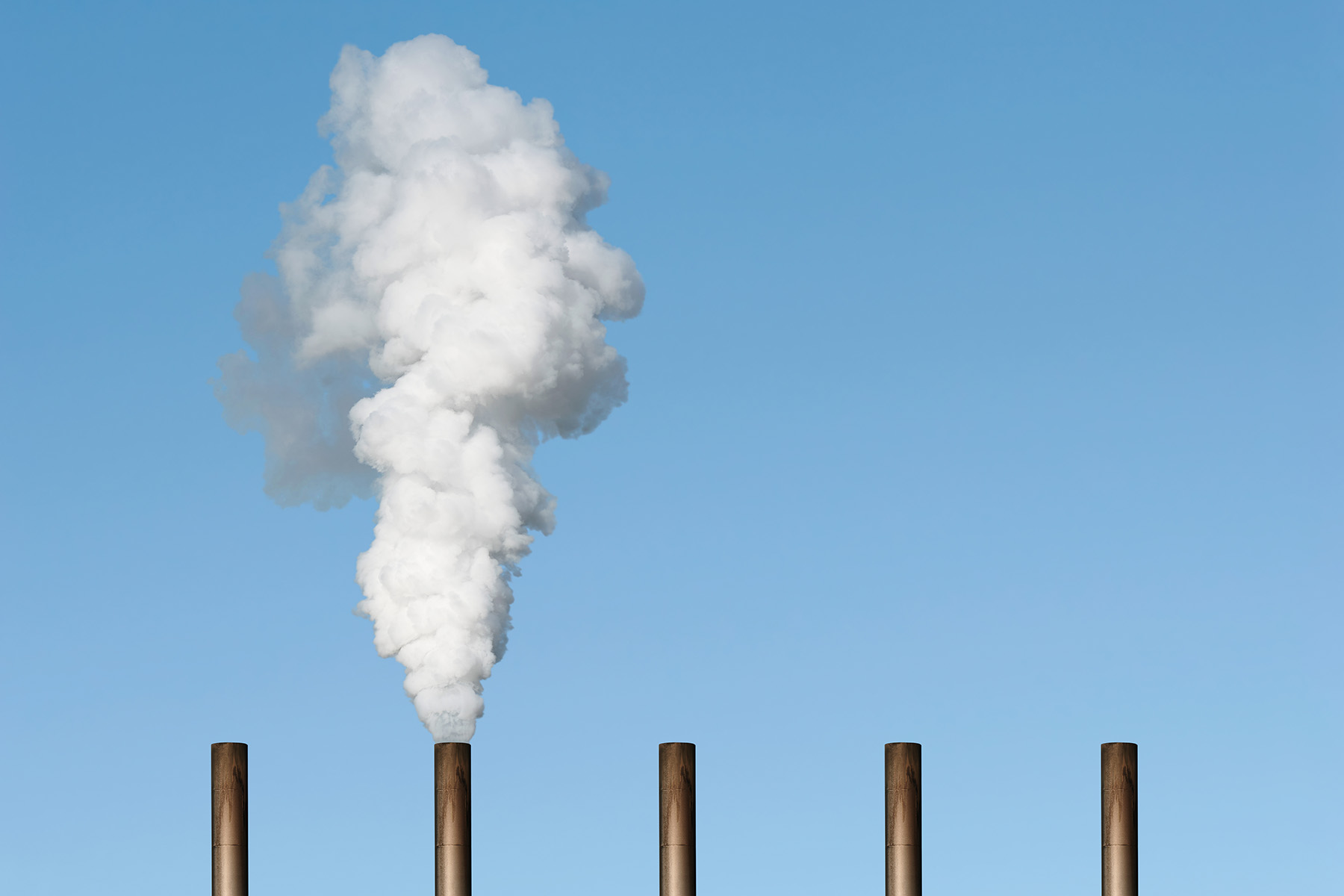 A plume of white smoke erupts from one of five smokestacks in front of a blue sky.