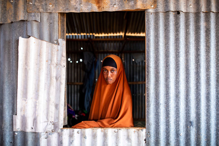 A woman in an orange hijab, staring out of an open window in a metal structure.