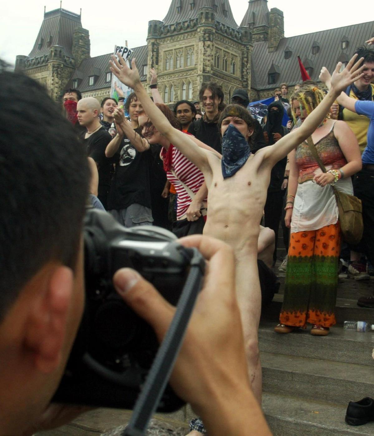 Photo of a naked man with his face covered by a bandana having his photo taken. He is standing in front of a crowd of protesters.