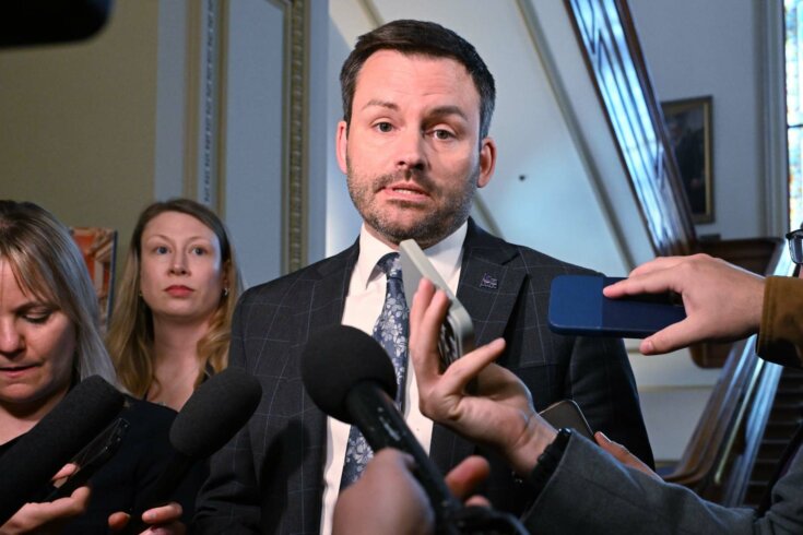 Photo of a man with short dark hair wearing a suit speaking to microphones while staring directly at the camera.