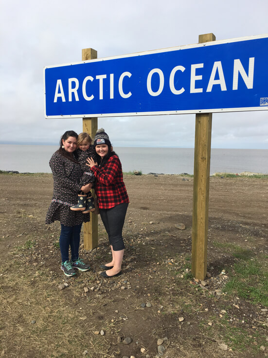 Two women and a toddler huddle together beneath a sign that reads "Arctic Ocean."