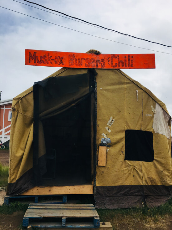 An open brown tent with a hand-drawn red sign that is advertising "Musk-ox burgers and chili."