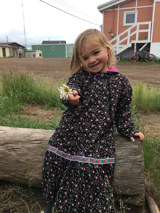 A toddler with long blonde hair in a floral printed dress holds a daisy to the camera and smiles.