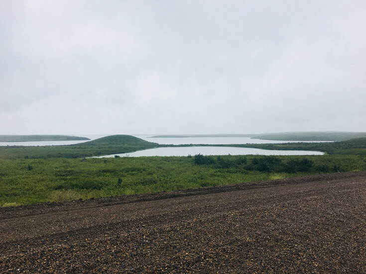 In the foreground is a dirt road; in the background are rolling green hills that contain small bodies of water.