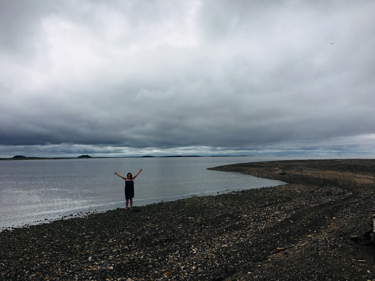 A woman stands on the rocky shore of a beach and raises her arms against a backdrop of water and a grey sky.