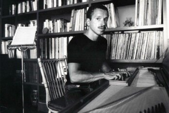 Black and white photo of a man with short hair and a moustache sitting at a piano in front of a wall of bookshelves.