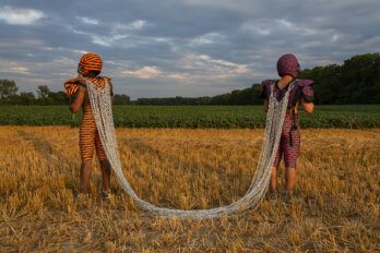 Two Black men stand in a harvested field under a cloudy sky. They are dressed in football uniforms printed with colourful African textiles. They are standing apart and facing away from each other, attached by a set of chains on their backs.