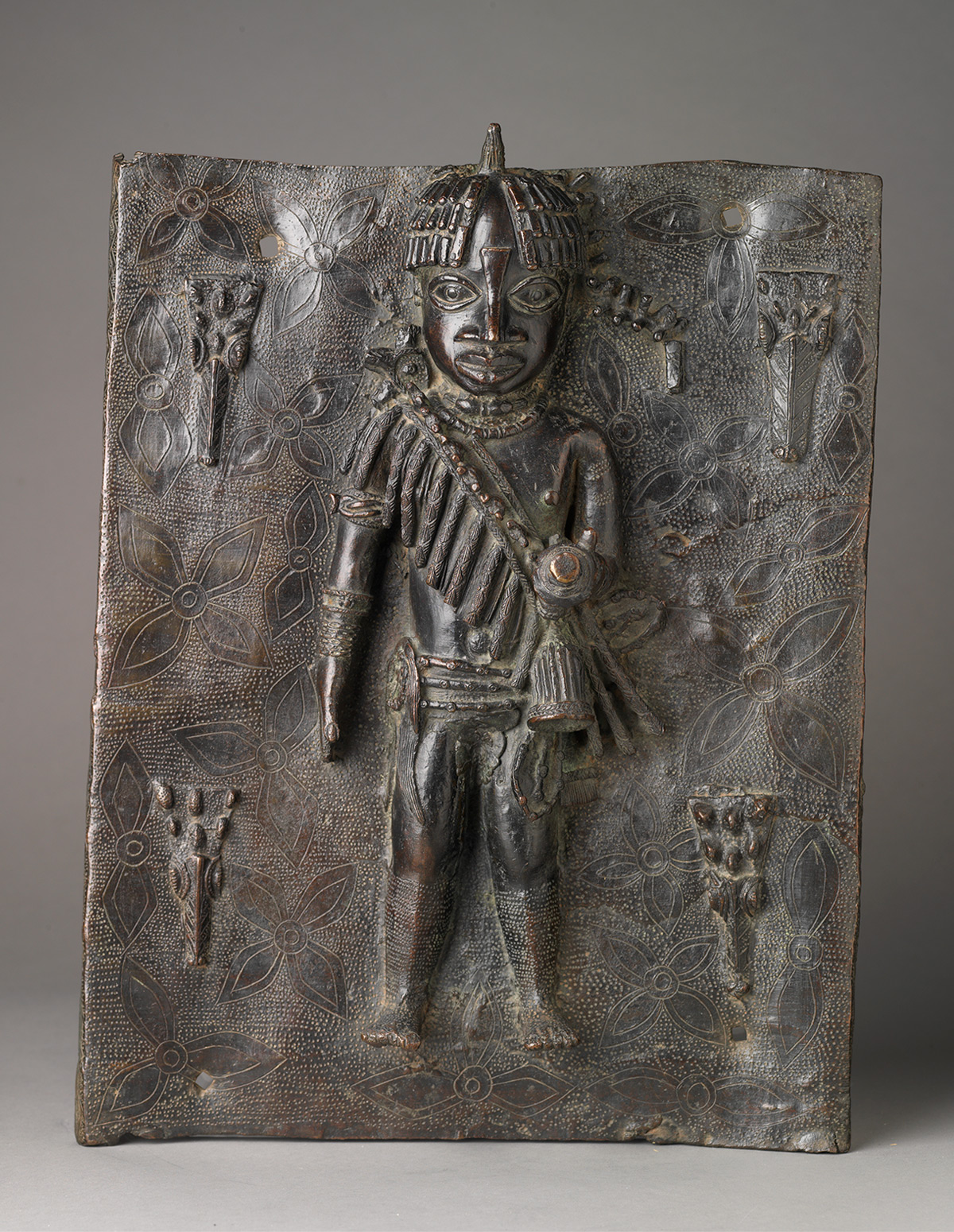 Photo of a rectangular metal plaque with central raised human figure on a background of dots and flowers.
