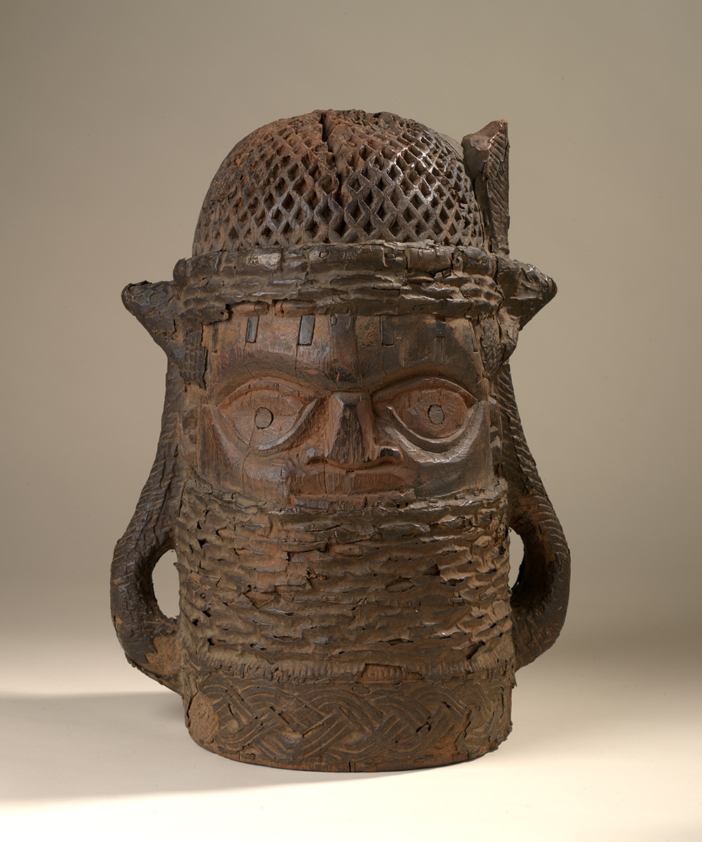 Photo of a wooden statue made to look like a head, cylindrical in shape with loops on each side representing braids of hair. It is wearing headgear with a honey-combed appearance. Brass overlays hide the figure’s chin and its eyes have wood inlays for the pupils.