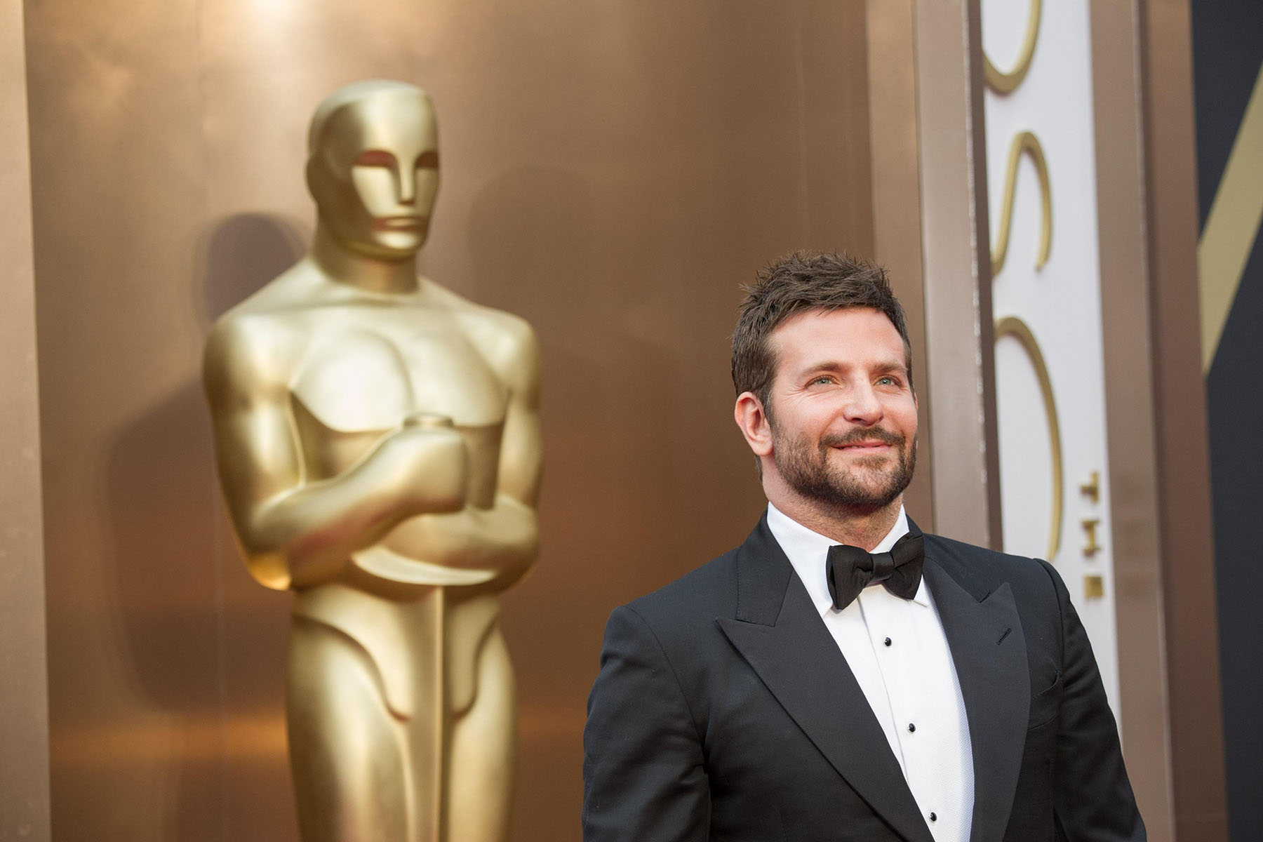 Bradley Cooper, dressed in a tux, smiles while a giant golden Oscar statute stands behind him.