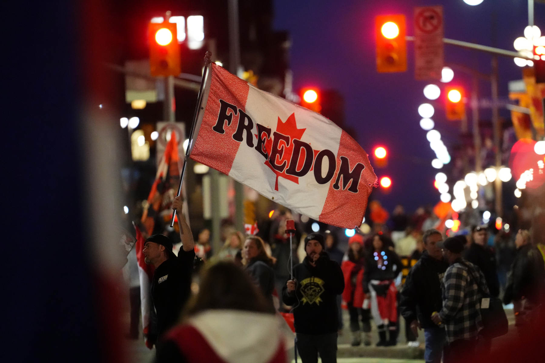 A protester waves a Canadian flag with 'freedom' written across it amid a nighttime protest.