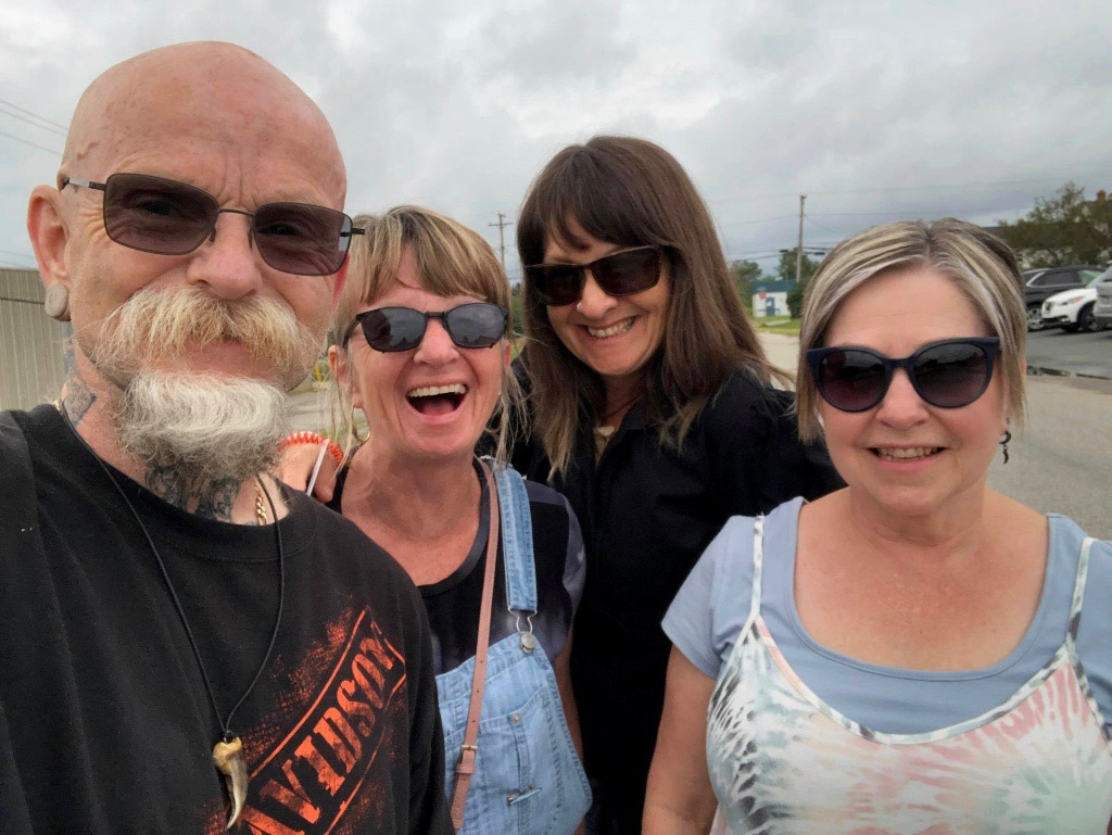 Photograph of a bald man with a beard and mustache standing beside three women. All are dressed casually and smiling at the camera.