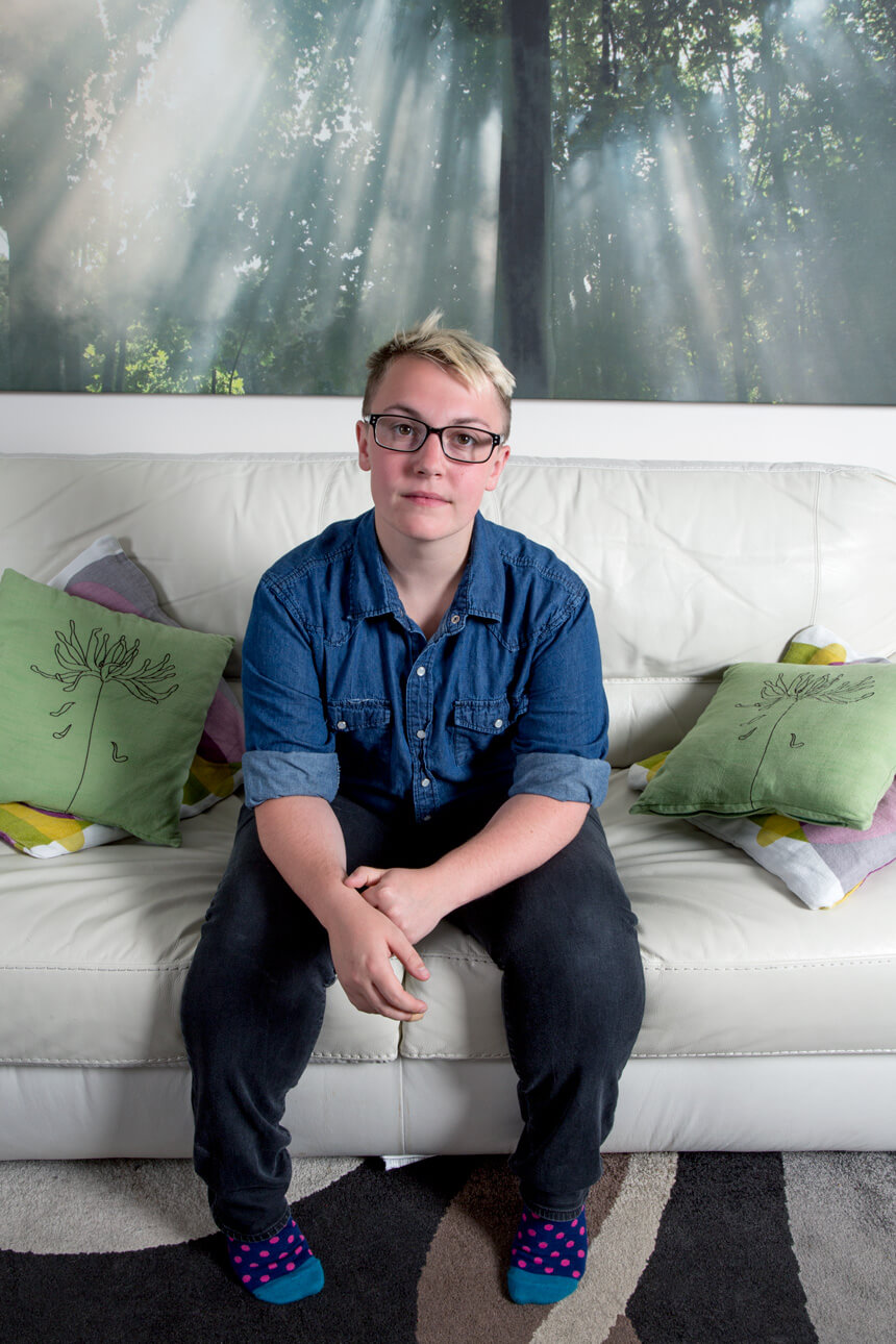 Photograph of a person with short blonde hair and thick-rimmed black glasses sitting on a couch.