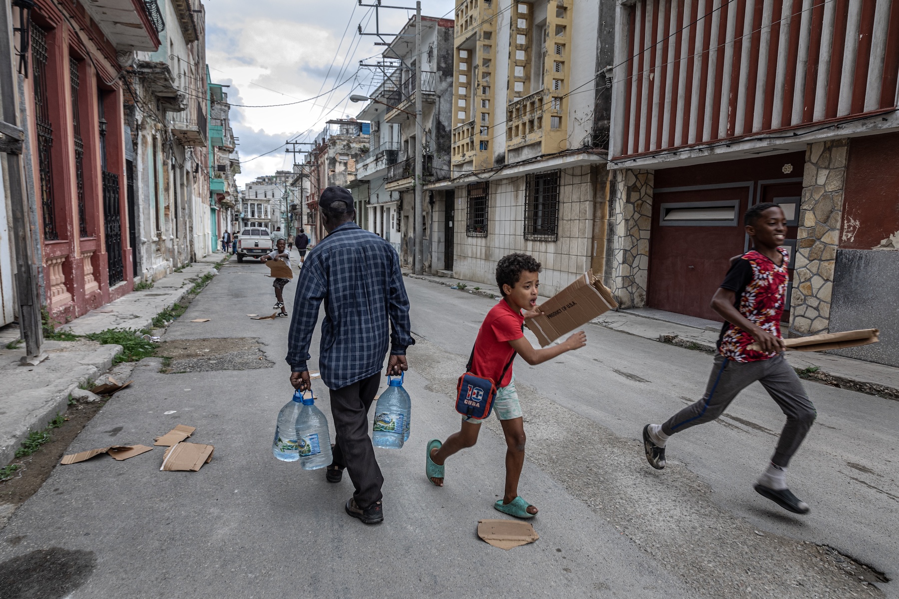 A photo of a person walking down the street with a jug of water. Two children are running with cardboard.