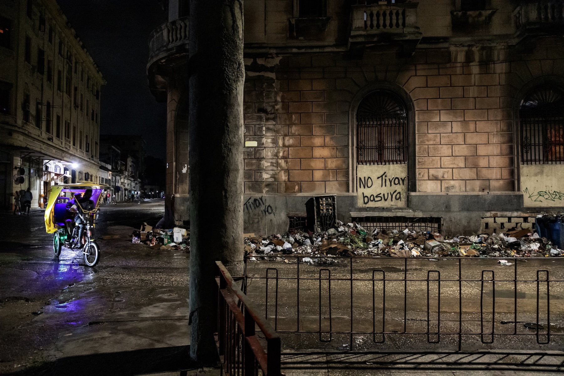 Photo of a brick building with trash piled up outside. A lighted bicycle vehicle is running down the street on the left side of the photo.